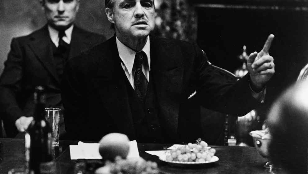 American actor Marlon Brando gestures at a table while American actor Robert Duvall sits behind him in a still from the film,