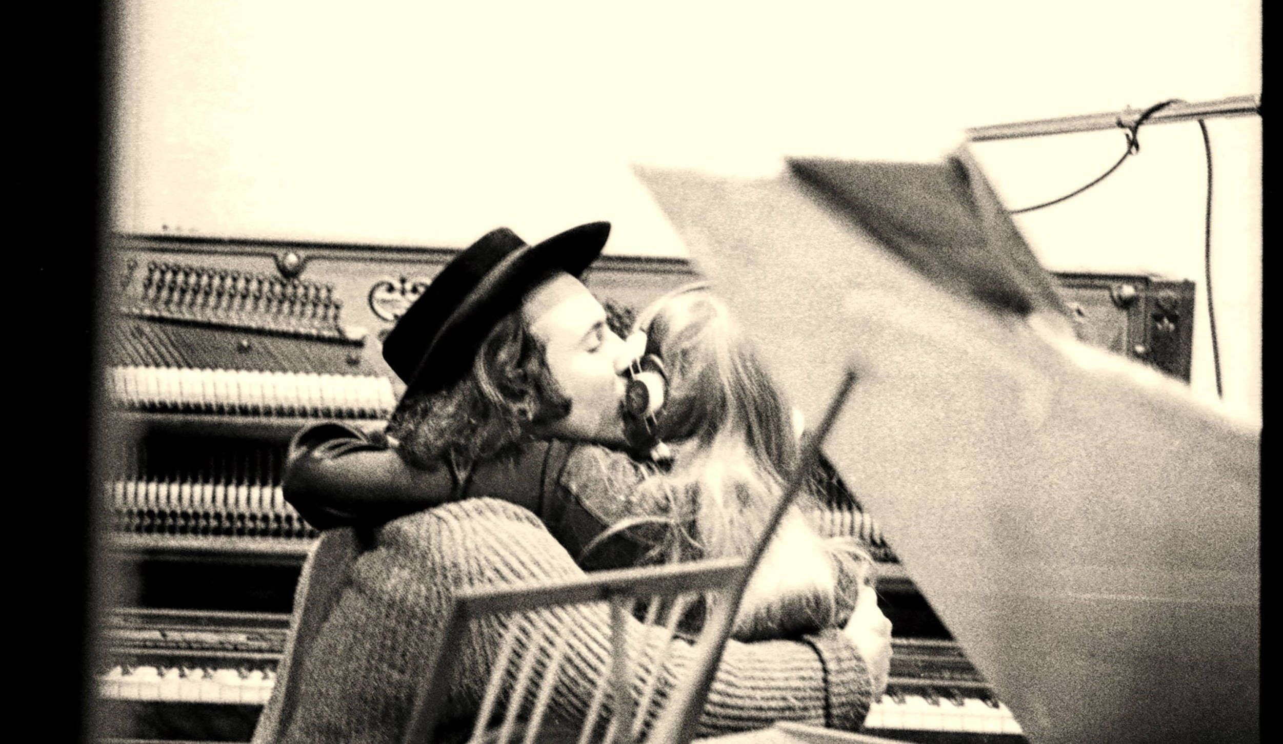 David Crosby kissing Joni Mitchell on the cheek while both are seated at an upright piano, seen from the back, through the...