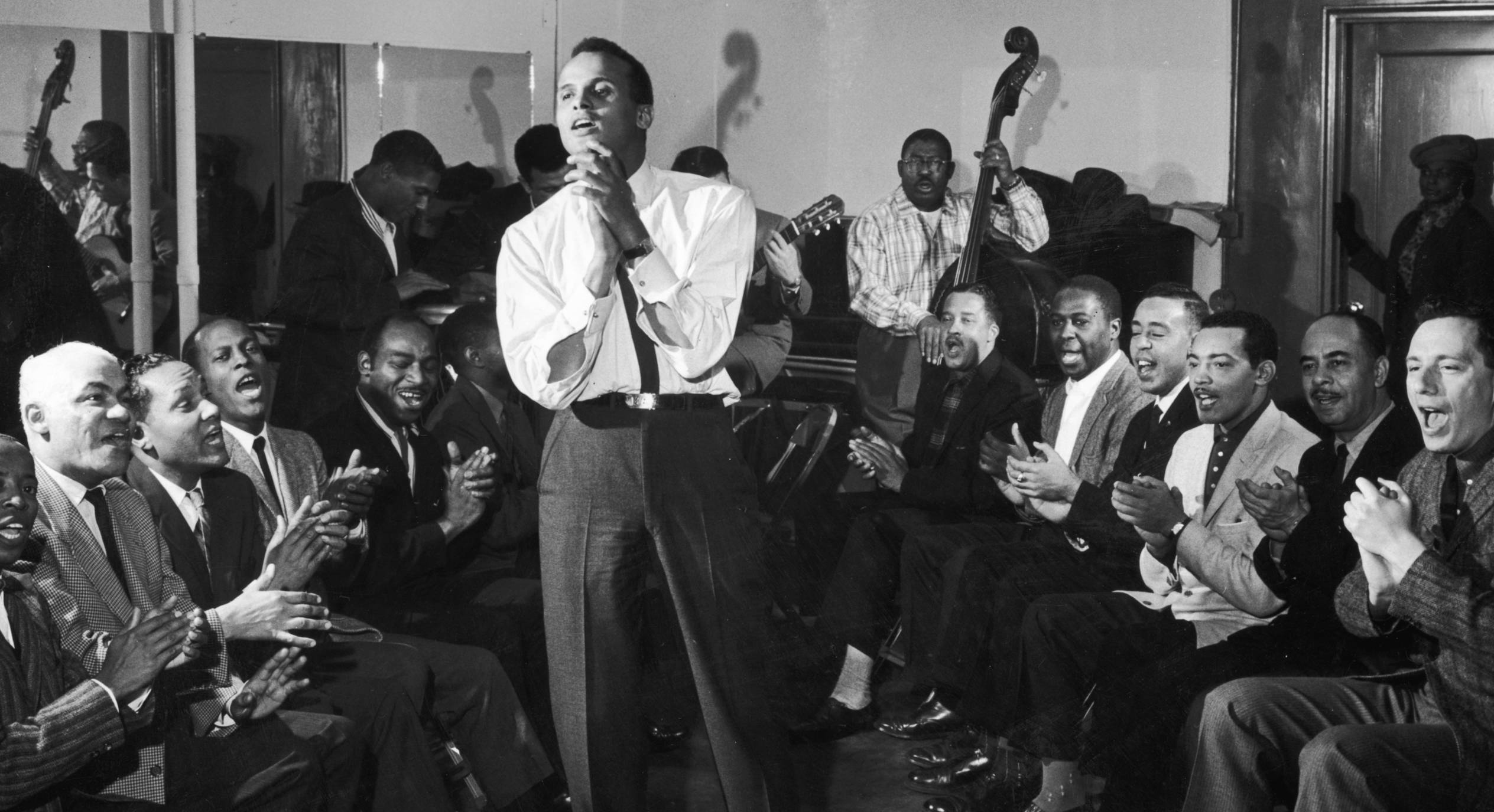circa 1955:  American singer and musician Harry Belafonte Jr. sings and claps his hands along with a group of men sitting ...