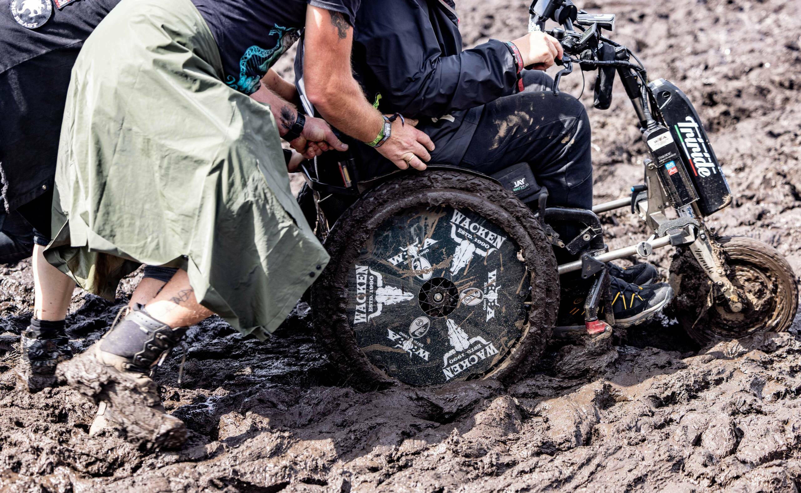 Ein Festivalbesucher hilft einer Person, die in einem Rollstuhl sitzt, sich ihren Weg durch den Schlamm auf dem Gelände d...