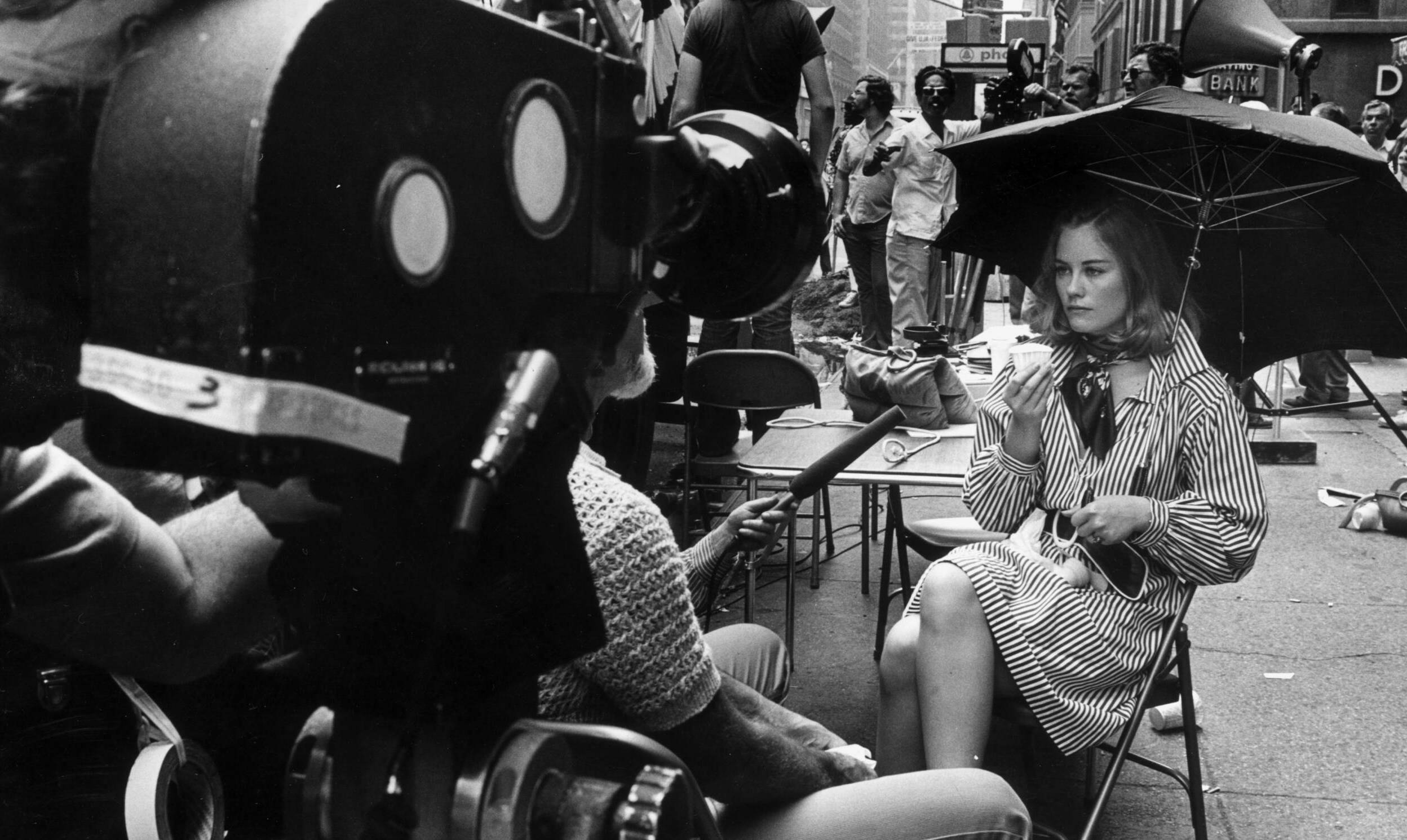 June 1975:  American actor Cybill Shepherd sits on the sidewalk with an umbrella and a cup, being interviewed, during the ...