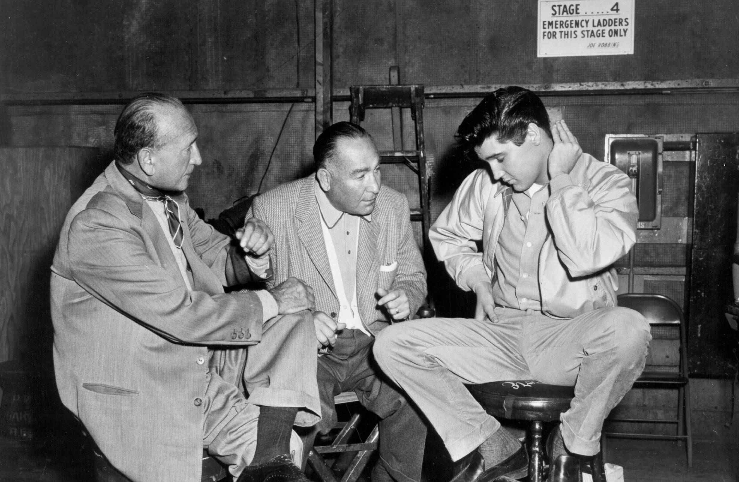 HOLLYWOOD - 1960: Rock and roll singer Elvis Presley confers with 2 men behind the scenes at a movie set talking to fans i...