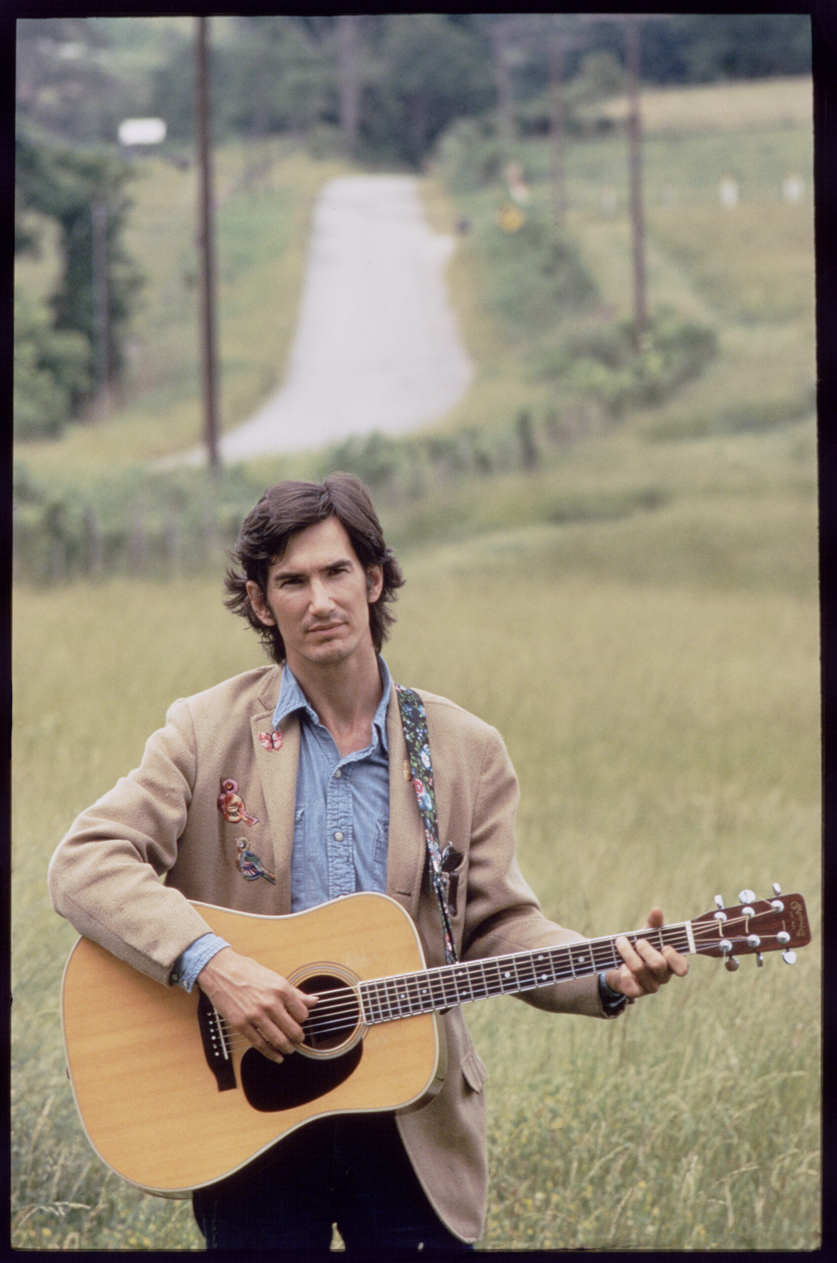 Singer, songwriter, musician, Townes Van Zandt shot waist up, standing in a field with winding road behind him, holding an...