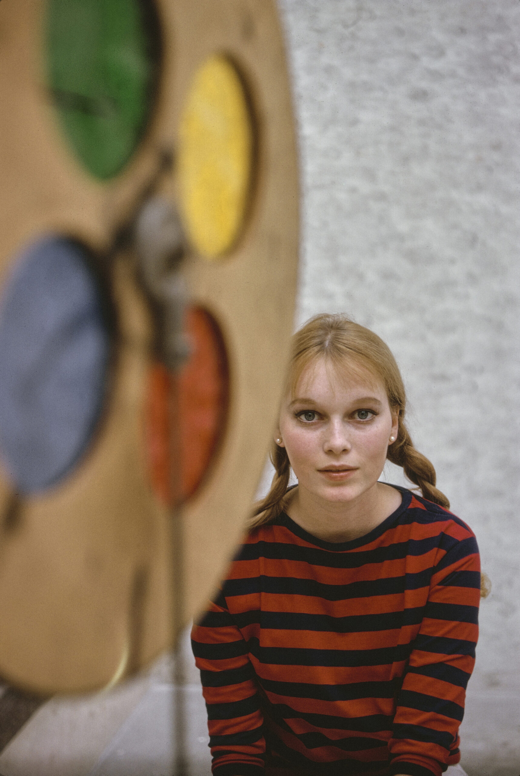 American actress Mia Farrow, wearing a red-and-black hooped long-sleeve outfit, with her hair in plaits, partially obscure...