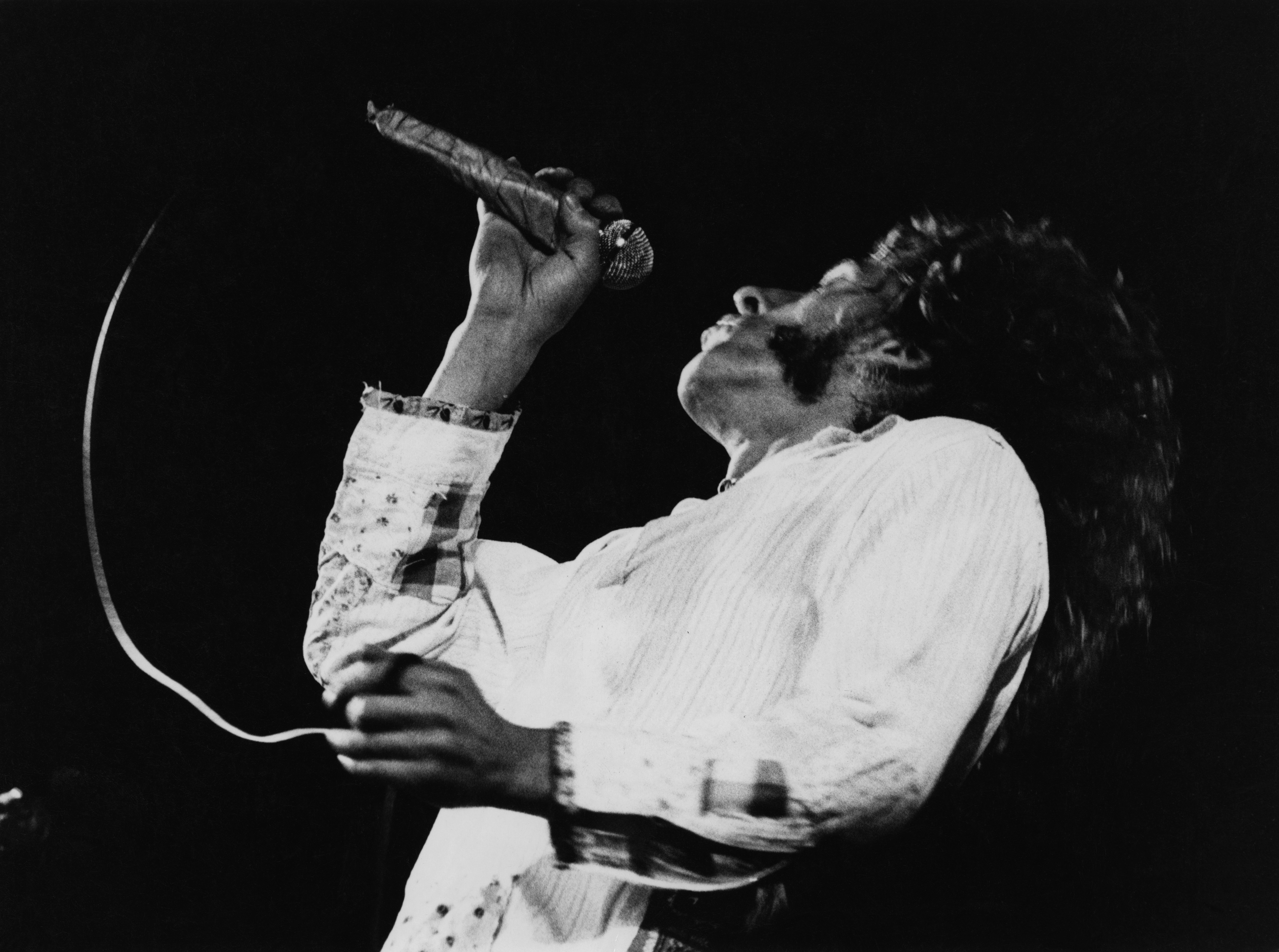 Roger Daltrey, singer with The Who, throwing his head back while singing into a microphone during a live concert performan...