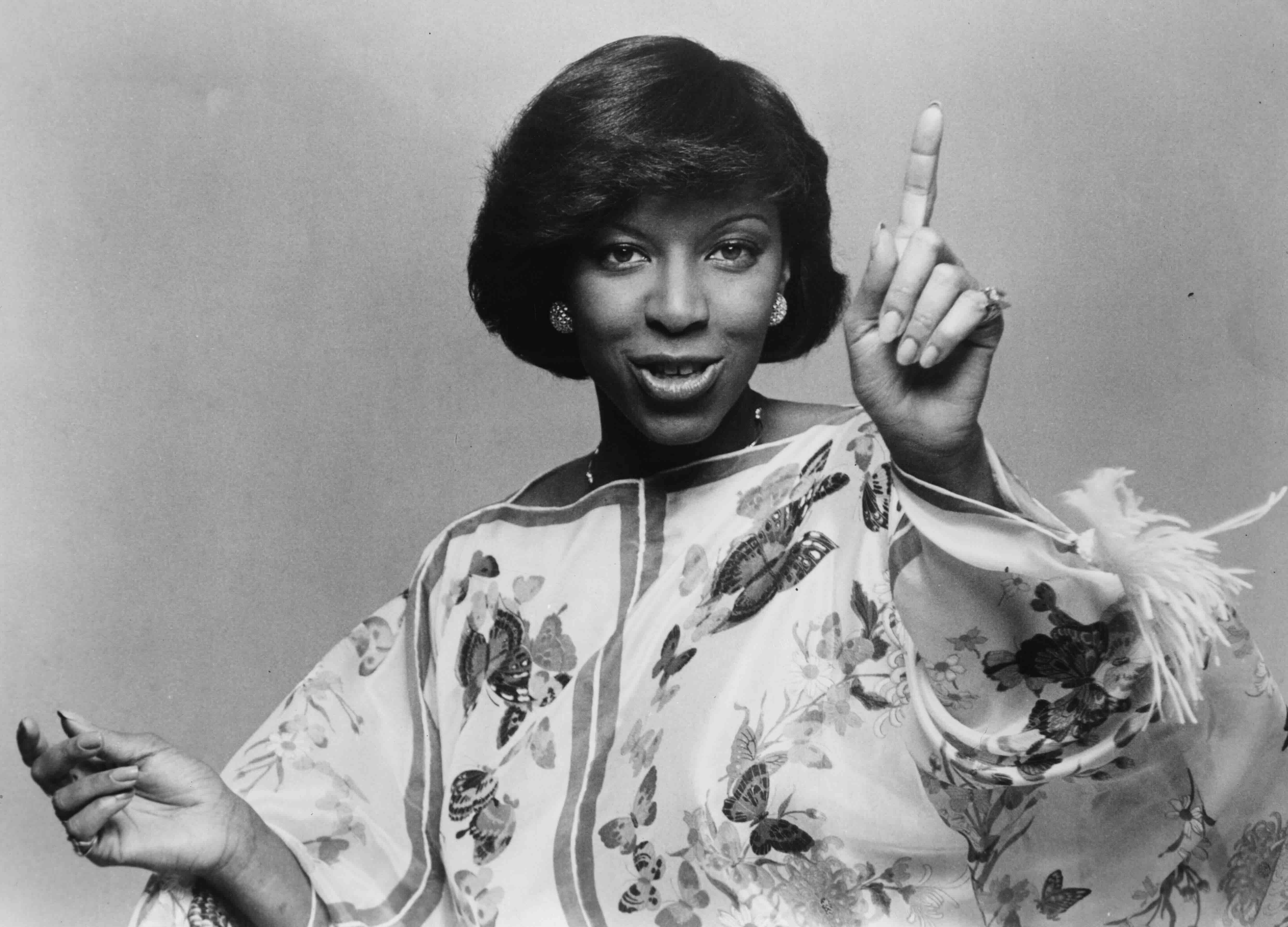 Portrait of singer Natalie Cole, pointing her finger and smiling, wearing a dress with a flower and butterfly pattern, cir...